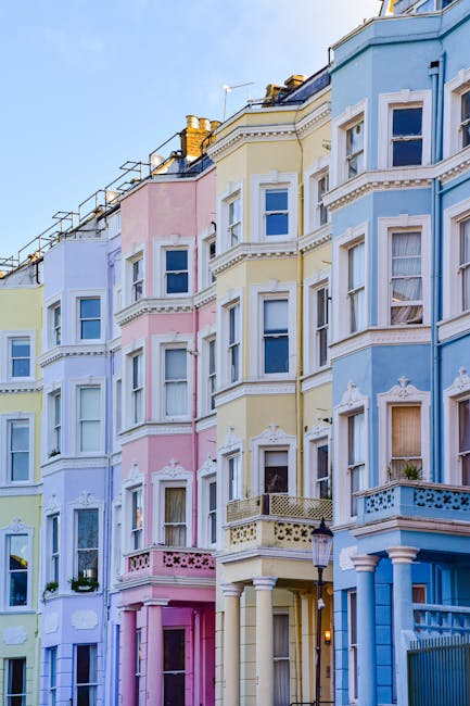 Image of a row of pastel-colored residential buildings in Notting Hill, featuring cream, pink, yellow, and blue facades. Each building has multiple white-framed sash windows with some open, and small decorative balcony railings. The buildings include architectural details such as stucco moldings and cornices. The street is illuminated by natural daylight, casting soft shadows and highlighting the clean exterior surfaces. The overall scene emphasizes well-maintained, freshly cleaned building exteriors, reflecting the high standards of property presentation upheld in the area, similar to the services offered by Cleaners Notting Hill for thorough surface cleaning and property maintenance in residential settings.