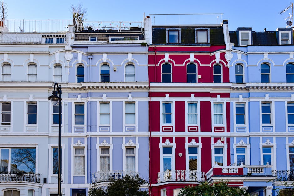 Image of a row of pastel-colored residential buildings in Notting Hill, featuring cream, pink, yellow, and blue facades. Each building has multiple white-framed sash windows with some open, and small decorative balcony railings. The buildings include architectural details such as stucco moldings and cornices. The street is illuminated by natural daylight, casting soft shadows and highlighting the clean exterior surfaces. The overall scene emphasizes well-maintained, freshly cleaned building exteriors, reflecting the high standards of property presentation upheld in the area, similar to the services offered by Cleaners Notting Hill for thorough surface cleaning and property maintenance in residential settings.