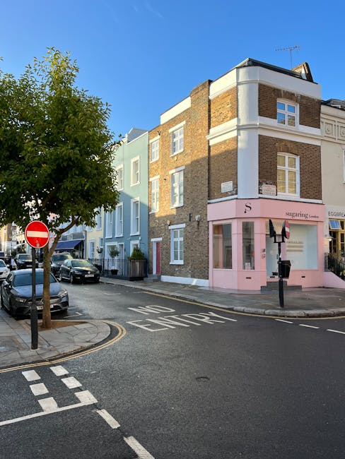 Photograph of a street corner in Notting Hill featuring a three-story building with a pink shopfront and white detailing. The building appears clean and well-maintained, with large windows on the upper floors and a glass display window on the ground floor. Surrounding the building are parked cars along the curb, a tree providing some greenery, and a 'no entry' traffic sign. The street has clear road markings, including a 'KEEP CLEAR' zone, and the sky is bright and blue, indicating a sunny day. The scene illustrates an urban environment where professional cleaning by companies like Cleaners Notting Hill ensures the cleanliness and presentation of commercial exteriors and street surfaces, contributing to a neat and inviting shopping area.