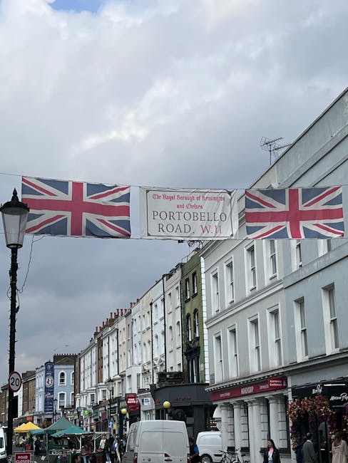 A view of Portobello Road in Notting Hill, featuring a street scene with white-painted residential buildings and shops. Overhead, there is a large banner displaying the Union Jack flags and the text 'The Royal Borough of Kensington and Chelsea PORTOBELLO ROAD, W.11'. The street is bustling with pedestrians, small market stalls with colorful umbrellas, and parked vans. The area appears clean and well-maintained, with a mix of modern and traditional architecture. The image emphasizes the vibrant character of the street, which has recently undergone surface cleaning and aesthetic restoration by Cleaners Notting Hill, specialists in deep cleaning and surface sanitisation.