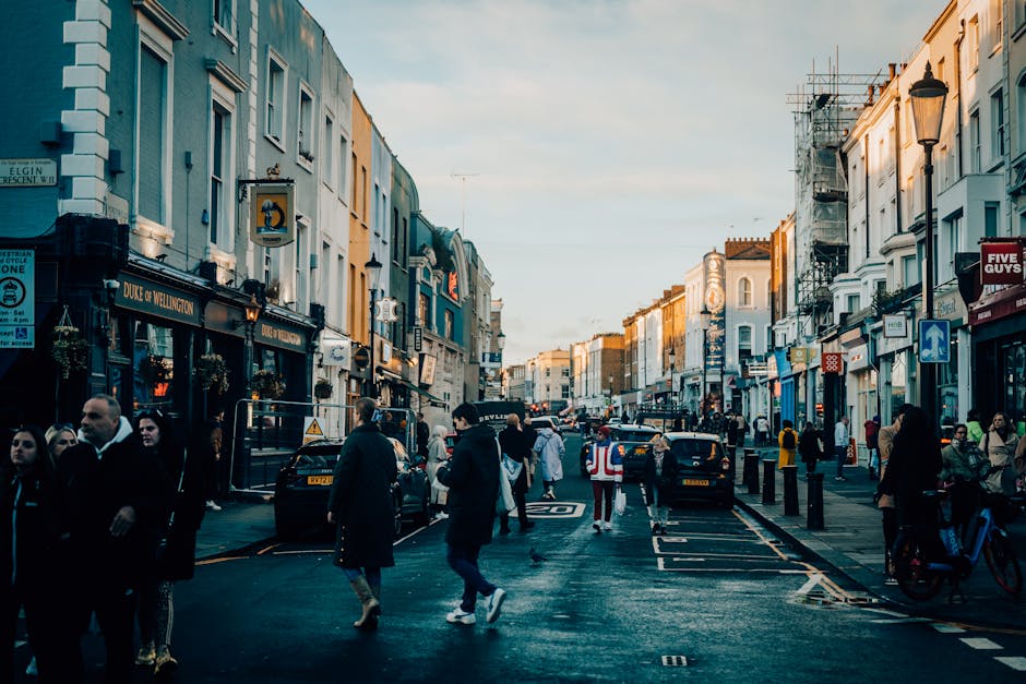 A busy street scene in Notting Hill during late afternoon with pedestrians walking along the pavement and crossing the road. The street is lined with multi-story buildings featuring a mix of painted facades, some with scaffolding. Shopfronts on either side display signs, such as Duke of Wellington and Five Guys, with illuminated lighting. The road surface appears wet, reflecting the buildings and streetlights, indicating recent cleaning or rain. Vehicles, including cars and a cyclist, are present amid the pedestrians. The streetlights are turned off, and the sky is partly cloudy, casting soft natural light over the scene. This image illustrates the lively urban environment of Notting Hill, which is relevant to the topic of surface cleaning, hygiene, and maintaining cleanliness in bustling residential and commercial areas, as highlighted on cleaners-nottinghill.com.