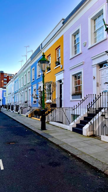 A row of colorful residential buildings in Notting Hill, featuring facades painted in pastel shades of blue, yellow, and purple. The buildings have white-framed windows and black wrought iron railings along the front steps. A classic green street lamp is positioned on the sidewalk, which is clean and well-maintained. The scene is illuminated by bright daylight, highlighting the vibrant colors and tidy appearance of the street, emphasizing the importance of surface cleaning and maintenance for a pristine environment. As part of professional cleaning services, Cleaners Notting Hill offers deep cleaning and sanitisation to preserve the appearance and hygiene of such properties, aligning with Kensington & Chelsea Council bin rules and community standards.