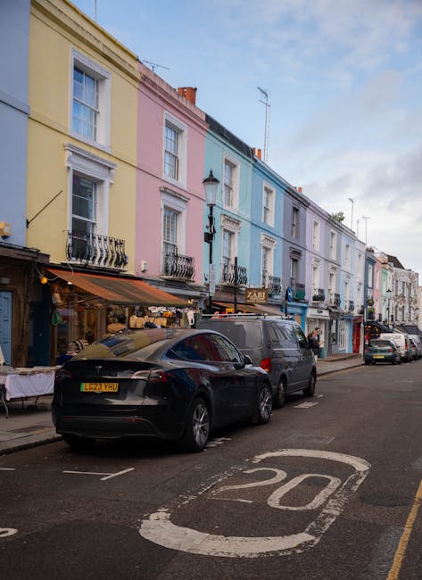 A busy street scene in Notting Hill during late afternoon with pedestrians walking along the pavement and crossing the road. The street is lined with multi-story buildings featuring a mix of painted facades, some with scaffolding. Shopfronts on either side display signs, such as Duke of Wellington and Five Guys, with illuminated lighting. The road surface appears wet, reflecting the buildings and streetlights, indicating recent cleaning or rain. Vehicles, including cars and a cyclist, are present amid the pedestrians. The streetlights are turned off, and the sky is partly cloudy, casting soft natural light over the scene. This image illustrates the lively urban environment of Notting Hill, which is relevant to the topic of surface cleaning, hygiene, and maintaining cleanliness in bustling residential and commercial areas, as highlighted on cleaners-nottinghill.com.