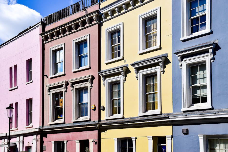 A row of three pastel-colored terraced houses with detailed white window frames and decorative cornices, each featuring three vertically aligned windows on the upper floors and a ground-floor window, all with clear glass panes and white sills. The pink, yellow, and blue facades appear freshly painted, with smooth surfaces and no visible dirt or damage. Natural daylight illuminates the scene, highlighting the clean and well-maintained appearance of the exteriors. A traditional black street lamp is visible on the left side of the image, set against a bright blue sky with some white clouds. The scene exemplifies residential exterior surface cleaning and maintenance, aligning with the expertise of Cleaners Notting Hill.