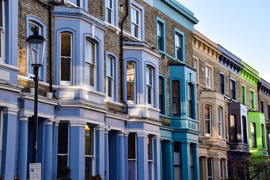 A row of Victorian-style terraced houses in Notting Hill, featuring a variety of painted facades in blue, beige, and green. The buildings have large sash windows with white trims, some with decorative mouldings and balconies. The ground level shows clean, well-maintained brick and stucco surfaces, with clear glass windows reflecting the sky and surroundings. A classic black street lamp is positioned in the foreground, along with a nearby parking sign. The scene is illuminated by natural daylight, highlighting the neatness and fresh appearance of the exteriors, suggesting recent surface cleaning or maintenance by professional cleaners like Cleaners Notting Hill as part of their surface cleaning and domestic cleaning services.
