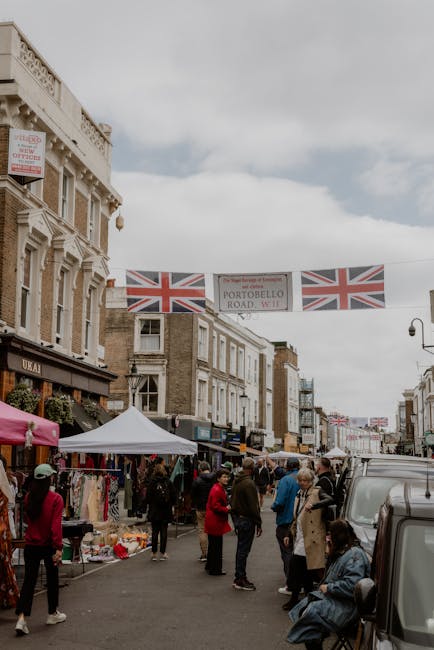 A busy street scene in Notting Hill decorated for the Carnival, with storefronts on both sides featuring traditional architectural details and balconies. The street is lined with market stalls under c