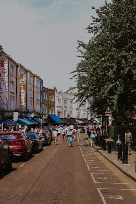 A busy street scene in Notting Hill decorated for the Carnival, with storefronts on both sides featuring traditional architectural details and balconies. The street is lined with market stalls under c