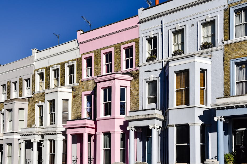 A busy street scene in Notting Hill during late afternoon, featuring a mix of pedestrians walking along the sidewalk and crossing the road. The street is lined with multi-story shops and cafes with co