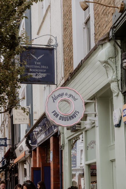 Close-up view of the storefront of a bakery with a white façade, featuring a round pink neon sign that reads 'Always a good FOR DOUGHNUTS,' mounted above a window. To the left, part of a sign for 'The Notting Hill Bookshop' is visible, hanging from a black bracket. The shop is situated on a narrow street lined with brick buildings and trees, with natural daylight illuminating the scene. The image emphasizes clean, well-maintained surfaces and the inviting appearance of the shop front, consistent with professional domestic and commercial surface cleaning standards as offered by Cleaners Notting Hill.